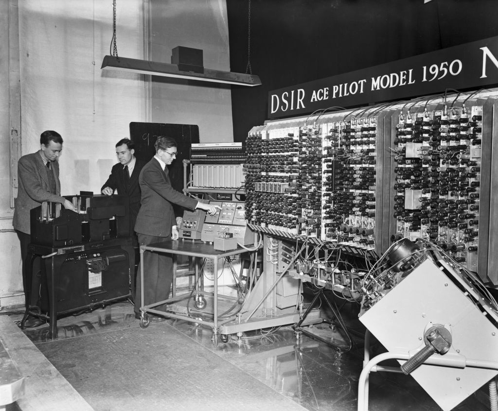 รุ่นเดอะ: แบบจำลองเครื่องคอมพิวเตอร์อัตโนมัติ (เอซ) รุ่นนำร่องแสดงต่อหน้าสื่อมวลชนในห้องปฏิบัติการฟิสิกส์แห่งชาติ ในกรุงลอนดอน สหราชอาณาจักร เมื่อวันที่ 29 พ.ย. 2483 วิศวกรอิเล็กทรอนิกส์ นายเอดเวิร์ด นิวแมน และนักคณิตศาสตร์ นายเจมส์ เอช วิลคินสัน ปรากฏอยู่ในภาพ 2 คนซ้ายจากทั้งหมด 3 คน ชิ้นส่วนคอมพิวเตอร์ ที่มีเครื่องแสดงผล แผงควบคุม และตัวเครื่องหลัก ก็ปรากฏต่อหน้าสื่อมวลชนเช่นกัน เอซเป็นคอมพิวเตอร์เครื่องแรกๆ ที่สร้างขึ้นในสหราชอาณาจักร และเป็นคอมพิวเตอร์ที่ติดตั้งโปรแกรมไว้ด้านใน ใช้หลอดสุญญากาศ 800 หลอด และหน่วยความจำที่ประกอบด้วยสายหน่วงปรอท ผู้ออกแบบ คือ นายอะลัน ทัวริง ก่อนลาออกจากห้องปฏิบัติการฟิสิกส์แห่งชาติไปเมื่อปี 2490